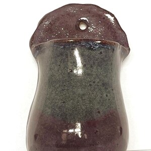 Może przedstawiać: Ścienny ceramiczny kwietnik z błyszczącym wykończeniem, posiadający głęboki śliwkowo-fioletowy dół i górę, kontrastujący z teksturowanym, nakrapianym ciemnoszarym środkiem oraz okrągłym otworem w górnej części do zawieszenia.