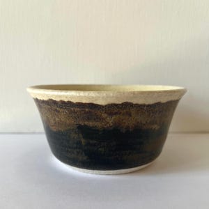 Könnte beinhalten: Eine kleine, weiße Keramikschüssel mit brauner und schwarzer Glasur. Die Schüssel hat eine abgerundete Form und einen leicht ausgestellten Rand.