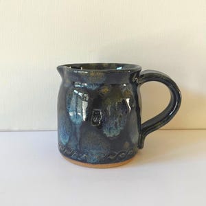 May include: A handmade ceramic pitcher with a dark blue glaze, accented with lighter blue and gold hues. The pitcher has a rounded body, a curved handle, and a small spout. The base is unglazed, revealing the natural clay color.
