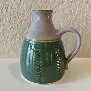May include: A small green ceramic pitcher with a light purple top and a handle. The pitcher has a fern design around the body.