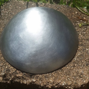 Puede incluir: Una cúpula de metal plateado, lisa y reflectante, se asienta sobre una superficie rugosa, gris y texturizada.
