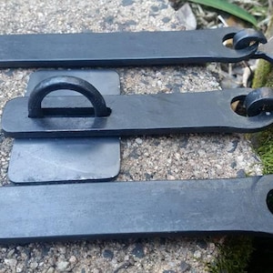 Puede incluir: Tres pestillos de puerta de metal negro con un asa de anillo y un gancho en cada extremo. Los pestillos están sobre una superficie de hormigón gris.