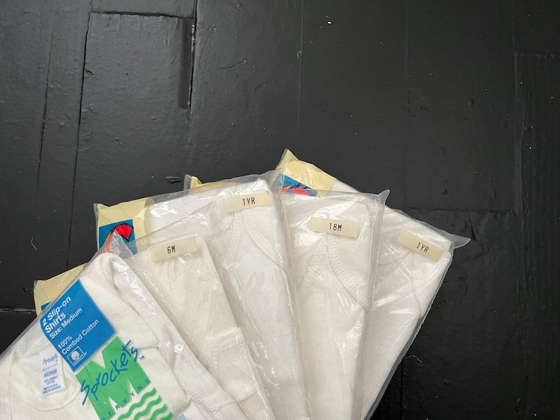 Puede incluir: Un paquete de cinco camisetas blancas para beb&eacute;s en diferentes tallas. Las tallas est&aacute;n etiquetadas como 6M, 1YR, 18M y 2YR. Las camisetas est&aacute;n hechas de 100% algod&oacute;n peinado.