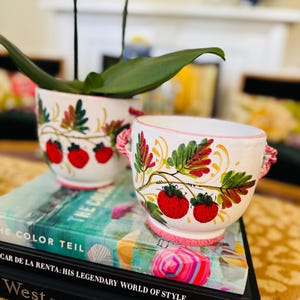 May include: Two white ceramic planters with hand-painted red strawberry motifs, green leaves, and pink rope-style handles. The planters feature a scalloped rim and a pedestal base, decorated with vibrant botanical illustrations of strawberries.