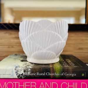 May include: A white ceramic planter featuring a scalloped, fan-like relief pattern across its exterior surface. The vessel has a rounded, bowl-shaped body with a smooth, glossy finish and a slightly flared rim, resting on a circular base.