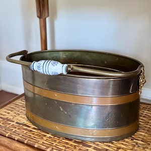May include: An oval brass bucket featuring two horizontal bands, a swing handle with a central white ceramic grip decorated with blue floral patterns, and small lion head accents on the sides where the handle attaches to the metal body.