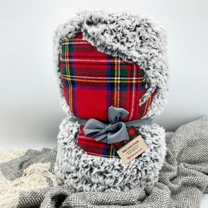 May include: A red and black plaid throw blanket with a gray faux fur trim. The blanket is tied with a gray ribbon and has a small tag that says "Fabi Loves Gracie" with a website address and social media handle.