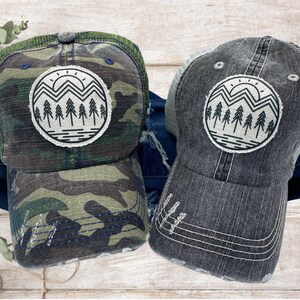 May include: A camouflage trucker hat and a gray trucker hat with a distressed look. The hat has a white and black graphic of a mountain range with a sun and trees.