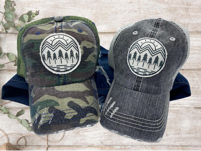 May include: A camouflage trucker hat and a gray trucker hat with a distressed look. The hat has a white and black graphic of a mountain range with a sun and trees.