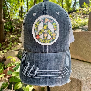 May include: A black denim trucker hat with a white mesh back. The hat features a large embroidered peace sign with colorful flowers around the edge.