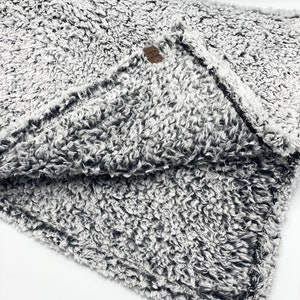 May include: A soft, gray and white fuzzy blanket with a black trim. The blanket is folded in half and the tag is visible.