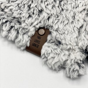 May include: A brown leather tag with the text "Fabi Loves Gracie" attached to a gray fuzzy blanket.