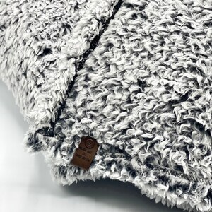 May include: A gray and white fluffy throw pillow with a brown leather tag that says "Machine Wash Cold Tumble Dry Low".