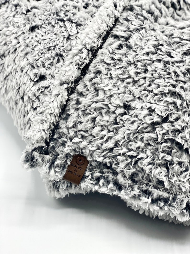 May include: A gray and white fluffy throw pillow with a brown leather tag that says "Machine Wash Cold Tumble Dry Low".