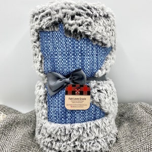 May include: A gray and blue blanket with a denim-like pattern. The blanket is rolled up and tied with a gray ribbon. A tag is attached to the ribbon with the text "Fabi Loves Gracie" and contact information.