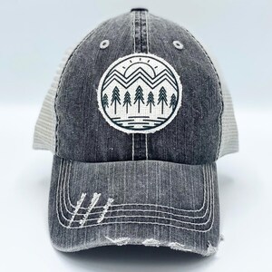 May include: A gray trucker hat with a distressed look. The hat has a mesh back and a black front with a white and black graphic of a mountain range with a sun and trees.