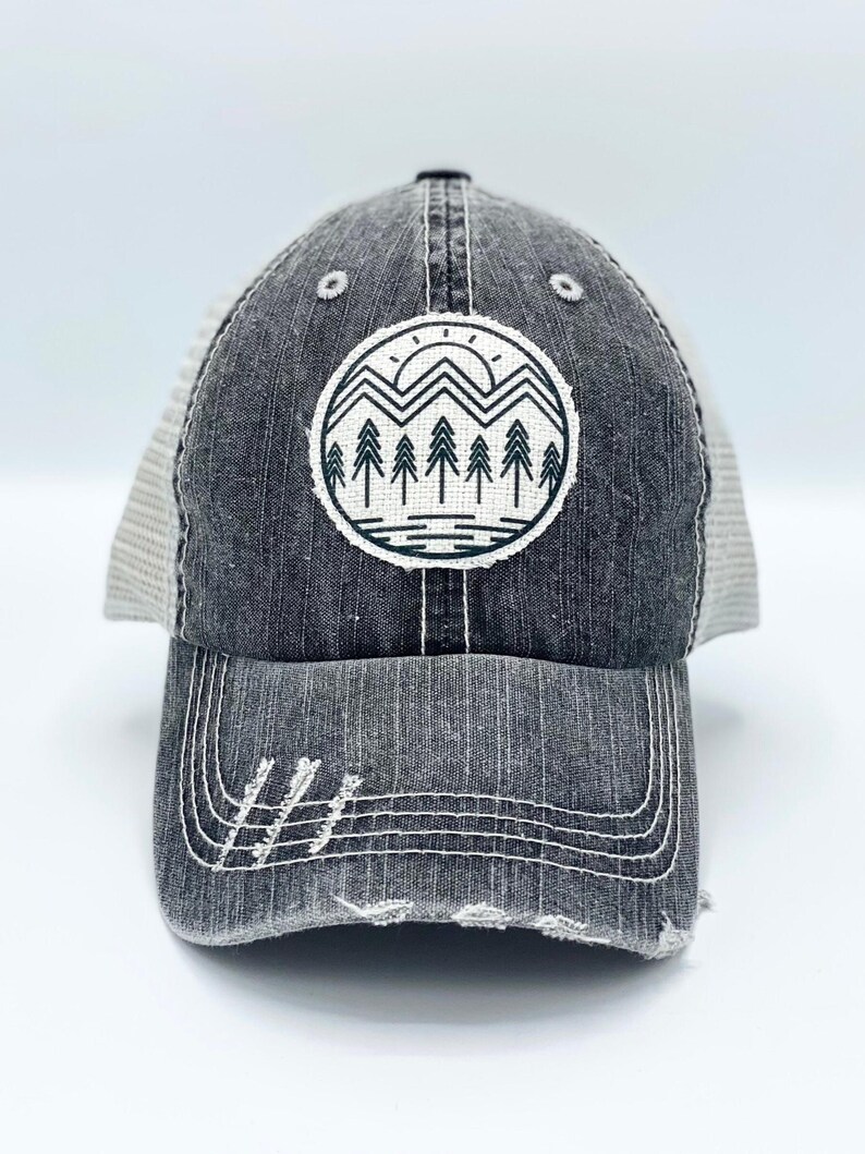 May include: A gray trucker hat with a distressed look. The hat has a mesh back and a black front with a white and black graphic of a mountain range with a sun and trees.