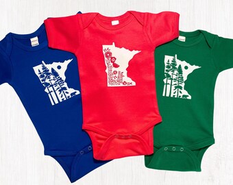 May include: Three baby onesies in blue, red, and green. Each onesie features a white graphic of the Minnesota state outline. The blue onesie has a forest design, the red has floral elements, and the green has a forest design.