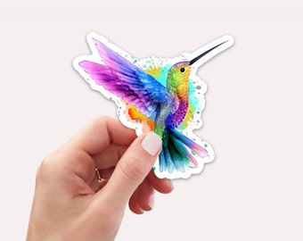 May include: A colorful hummingbird sticker with a watercolor effect. The hummingbird is flying with its wings spread and its beak open. The background is a light blue with splashes of yellow, orange, and green.