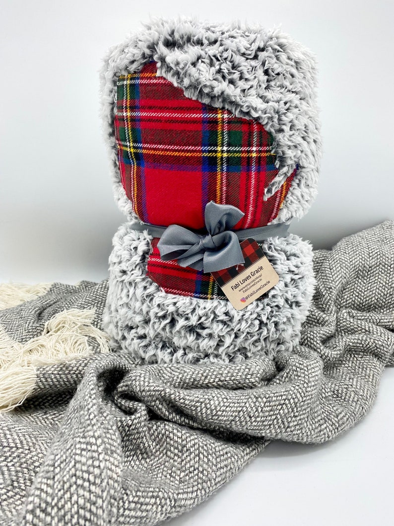 May include: A red and black plaid throw blanket with a gray faux fur trim. The blanket is tied with a gray ribbon and has a small tag that says "Fabi Loves Gracie" with a website address and social media handle.