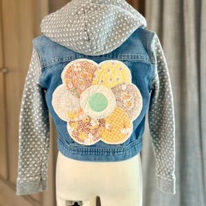 May include: A denim jacket with a gray hooded sweatshirt attached to the sleeves. The back of the jacket features a large, colorful patchwork flower.