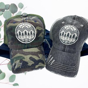 May include: A camouflage trucker hat and a gray trucker hat with a distressed look. The hat has a white and black graphic of a mountain range with a sun and trees.