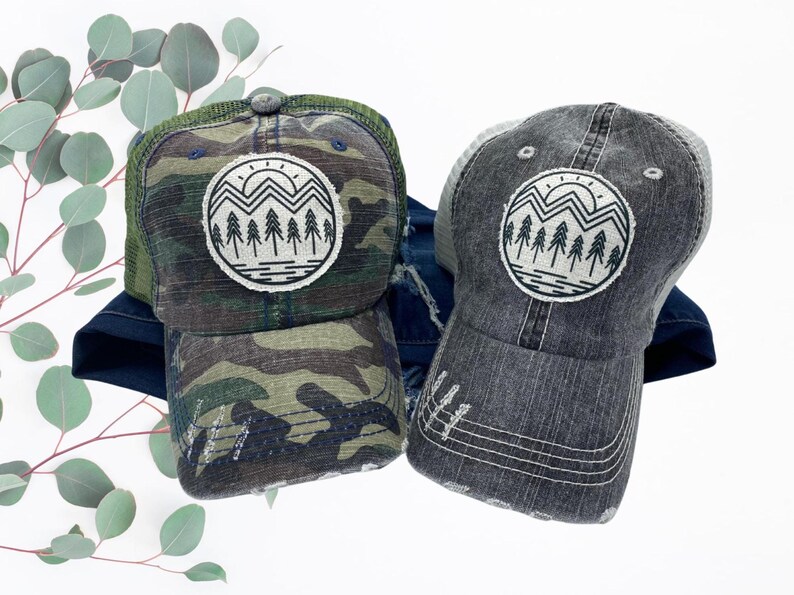 May include: A camouflage trucker hat and a gray trucker hat with a distressed look. The hat has a white and black graphic of a mountain range with a sun and trees.