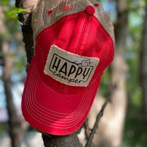 May include: Red and beige mesh trucker hat with a patch that says "Happy Camper" and a small graphic of a campfire.