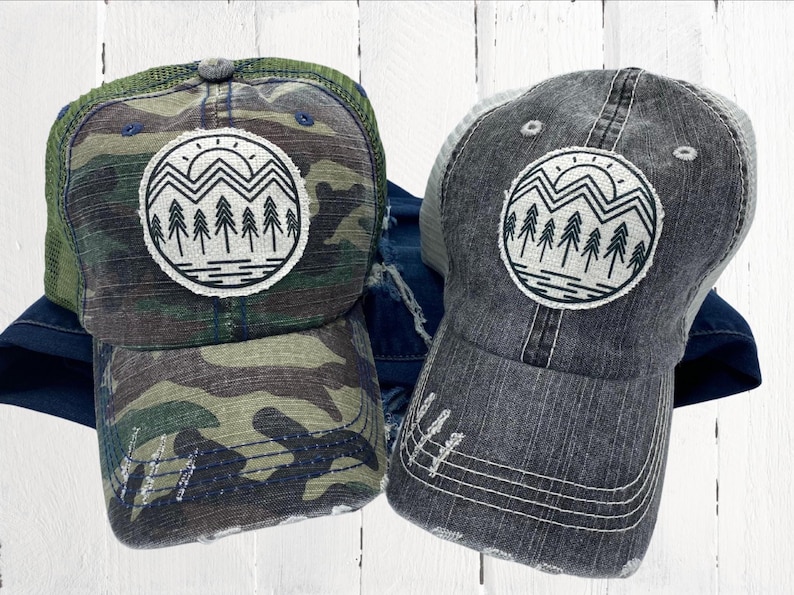 May include: A camouflage trucker hat and a gray trucker hat with a distressed look. The hat has a white and black graphic of a mountain range with a sun and trees.