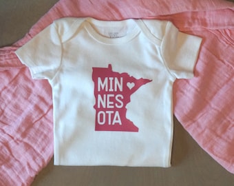 May include: White baby onesie with a pink outline of the state of Minnesota with a heart in the middle and the words "MINNESOTA" written vertically.