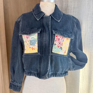 May include: A dark wash denim jacket with two front pockets featuring colorful patchwork designs. The jacket has a classic collar, a zipper closure, and a cropped fit. The patchwork includes floral and geometric patterns in various colors.
