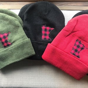 May include: Three knit beanies in different colors: green, black, and red. Each beanie features a black and red plaid outline of the state of Minnesota.