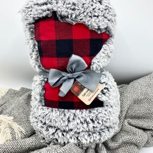 May include: A gray and white faux fur blanket with a red and black buffalo plaid fabric insert. The blanket is tied with a gray satin ribbon and has a small tag that says "Fabi Love" with a website address.
