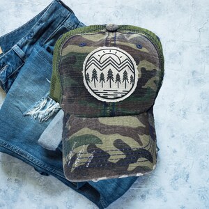 May include: A camouflage trucker hat with a distressed look. The hat has a mesh back and a black front with a white and black graphic of a mountain range with a sun and trees.