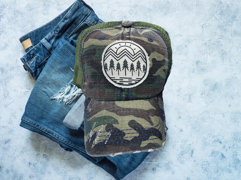 May include: A camouflage trucker hat with a distressed look. The hat has a mesh back and a black front with a white and black graphic of a mountain range with a sun and trees.
