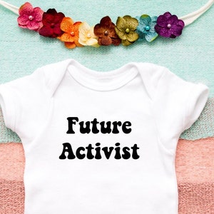 May include: White baby onesie with the text "Future Activist" in black, retro-style font. The onesie is lying on a multi-colored striped fabric background.