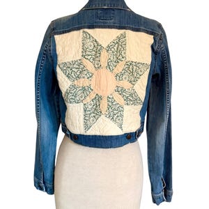 May include: A blue denim jacket featuring a large, square, quilted back panel. The panel displays a cream-colored fabric star with a central light pink circular patch and green floral patterned fabric accents within the star points.