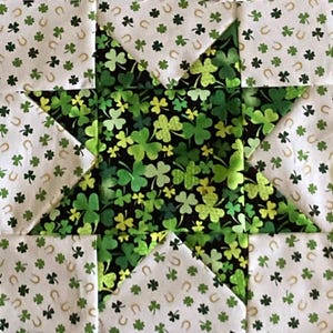 May include: A quilt block with a five-pointed star design. The star is made of black fabric with a green shamrock pattern. The background is white fabric with a green shamrock and gold horseshoe pattern.