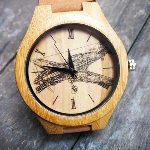 Puede incluir: Reloj de pulsera de madera con correa de cuero marrón claro. La esfera del reloj presenta una ilustración detallada de un avión. La caja del reloj es de madera clara. El reloj tiene agujas negras y números romanos.