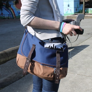 以下が含まれることがあります： フラップ開閉と調節可能な茶色のストラップが付いた、青と茶色のメッセンジャーバッグ。バッグのフロントポケットには、街並みのシルエットが描かれています。バッグは肩にかけて使用します。