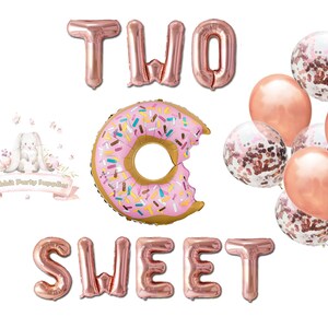 Op de afbeelding: Rosegouden folieballonnen met de tekst "TWO SWEET" en een donutvormige ballon met kleurrijke hagelslag. Er zijn ook verschillende roze en met confetti gevulde ballonnen, ideaal voor een tweede verjaardag.