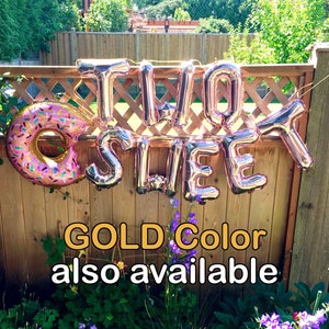 Op de afbeelding: Ros&eacute;gouden folieballonnen met de tekst "TWO SWEET" en een donutballon worden op een houten hek getoond. De donutballon heeft roze glazuur en kleurrijke hagelslag. De tekst hieronder luidt "GOLD Color also available".