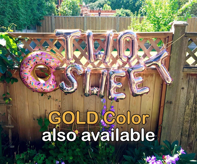 Op de afbeelding: Ros&eacute;gouden folieballonnen met de tekst "TWO SWEET" en een donutballon worden op een houten hek getoond. De donutballon heeft roze glazuur en kleurrijke hagelslag. De tekst hieronder luidt "GOLD Color also available".