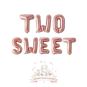 Op de afbeelding: Rosegouden folieballonnen met de tekst "TWO SWEET" op een witte achtergrond. De ballonnen zijn in twee rijen gerangschikt. Onderaan staat een logo van "Rabbit Party Supplies".