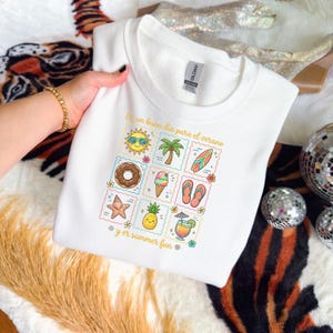 Puede incluir: Una sudadera blanca de cuello redondo con una cuadrícula de nueve ilustraciones veraniegas coloridas que incluyen un sol, una palmera, una tabla de surf, un dónut, un helado, chanclas, una estrella de mar, una piña y un cóctel, con el texto Es un buen día para el verano y er summer fun.