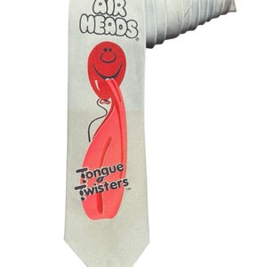 Puede incluir: Una corbata gris claro con el logotipo de Air Heads y un gráfico de lengua roja con las palabras "Tongue Twisters". La corbata está enrollada en la parte superior, mostrando su textura y diseño.