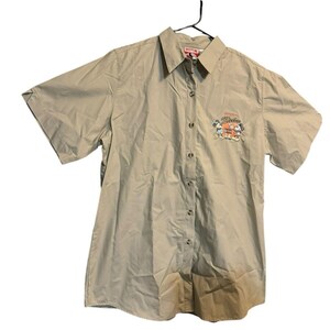 May include: Tan short-sleeve button-down shirt. The shirt features a small embroidered design of cartoon characters and the word "Mickey" in orange. The shirt is hanging on a black hanger.