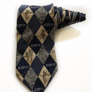 May include: A blue and tan patterned tie with the ESPN logo repeated throughout. The tie features a diamond pattern with images of golfers.