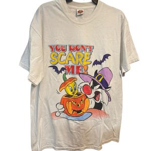 May include: White t-shirt featuring a Halloween-themed graphic. The design includes the text "YOU DON'T SCARE ME!" with cartoon characters Tweety Bird, Sylvester the Cat, and a jack-o'-lantern. The shirt has short sleeves and a crew neck.