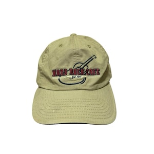 Puede incluir: Gorra de béisbol color beige con el logotipo de Hard Rock Cafe. El logotipo incluye una guitarra, las palabras "HARD ROCK CAFE" en rojo y un óvalo azul con "Est. 1971". La gorra tiene una visera curva y una correa de tela.